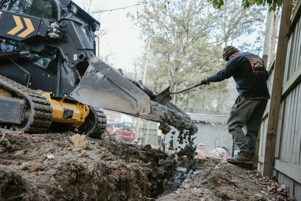 A worker shovelling material into a narrow backyard trench beside a compact loader
