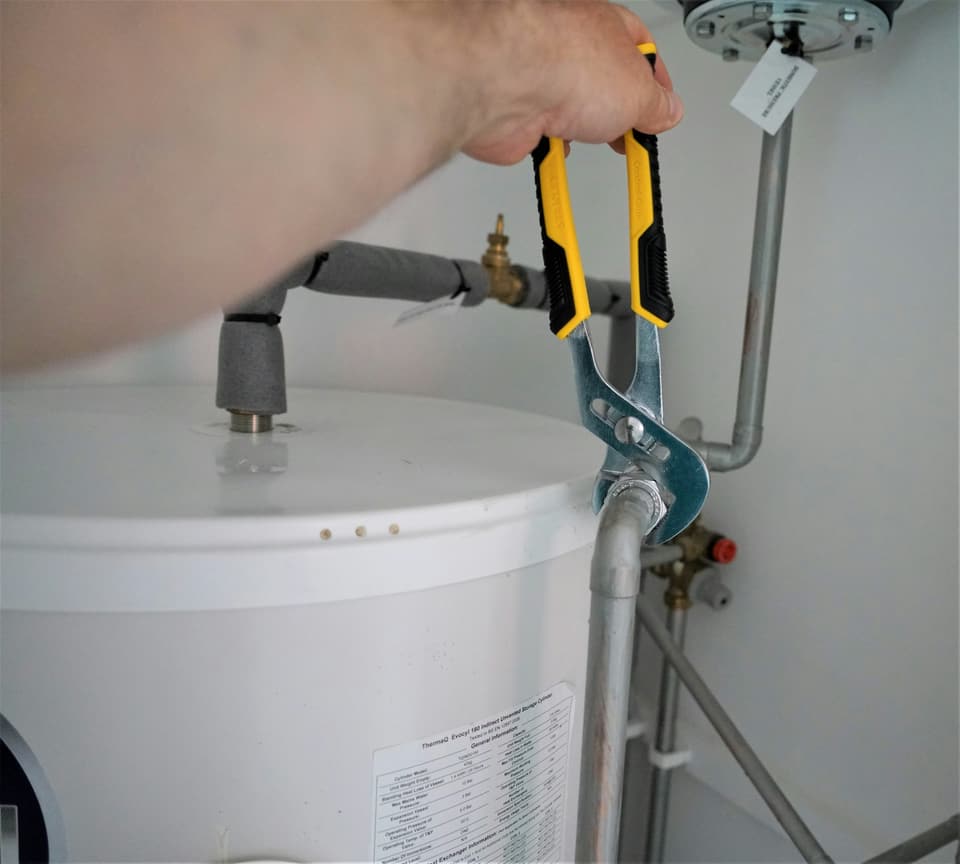 A hand using pliers on a pipe fitting at the top of a white hot water tank